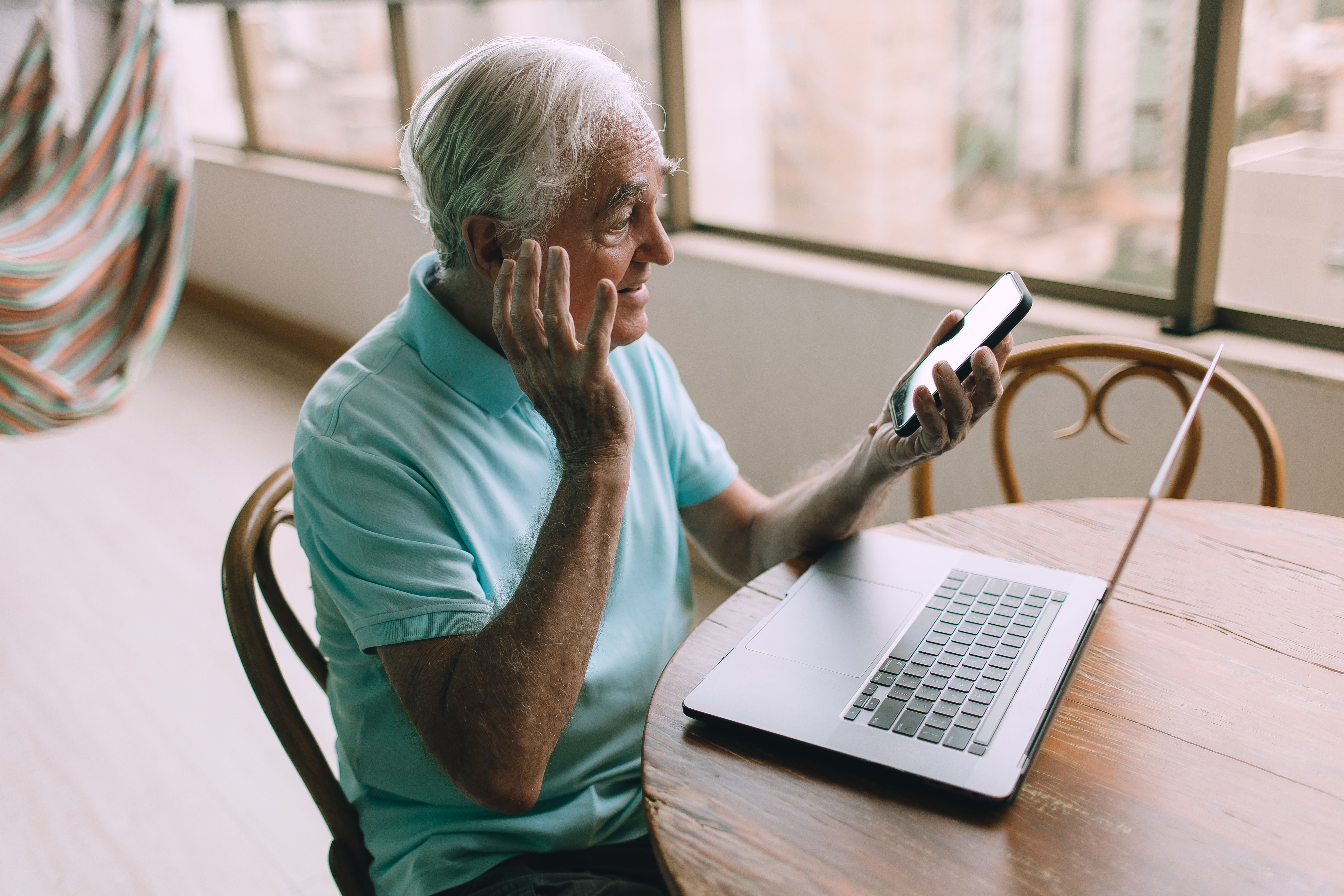 Homem idoso com camiseta azul usando smartphone e laptop em uma mesa de madeira dentro de casa.