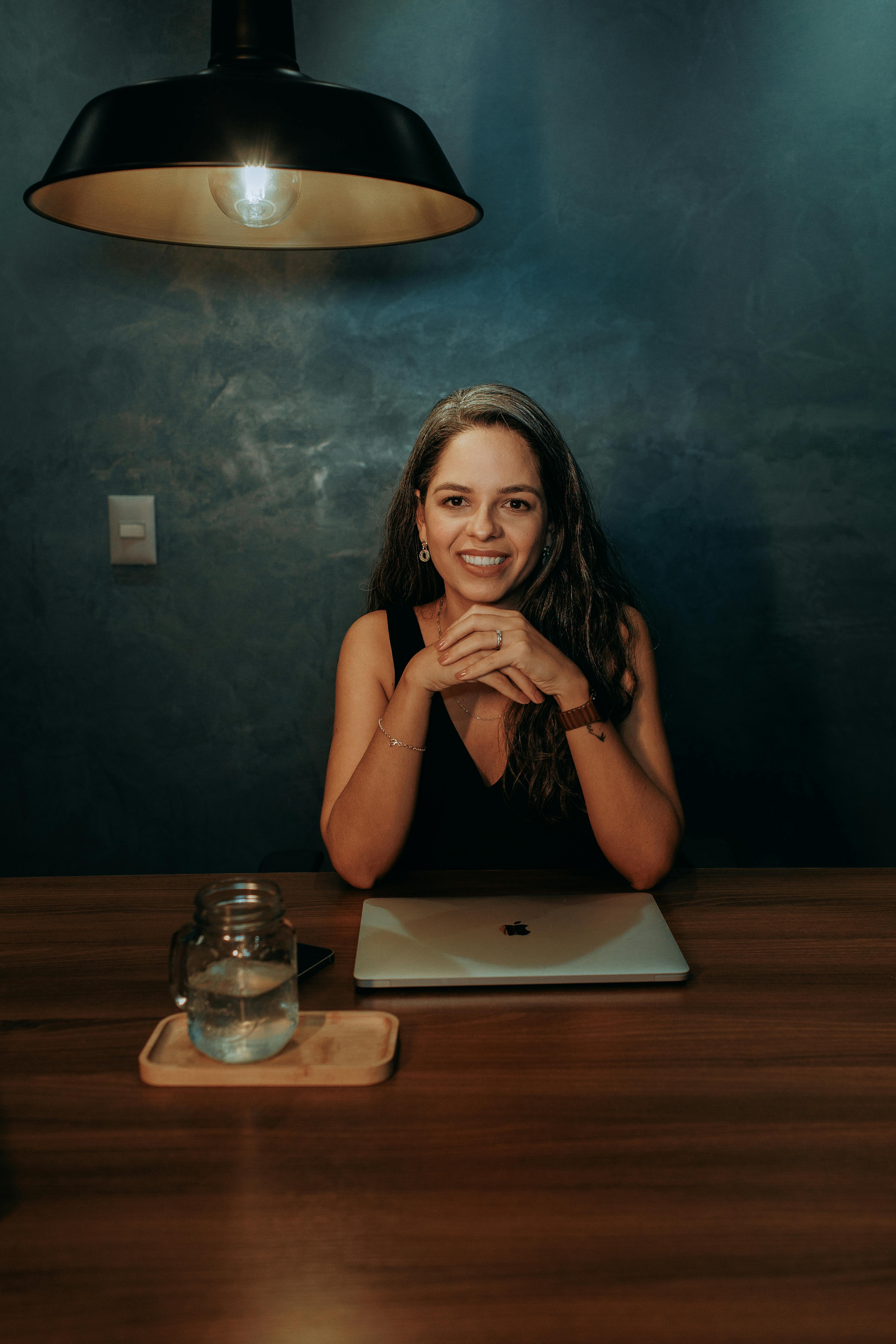 Ilmainen Hymyilevä nainen istuu työpöydän ääressä kannettava tietokoneensa kanssa lämpimässä sisävalaistuksessa, joka luo viihtyisän tunnelman. Kuvapankkikuva