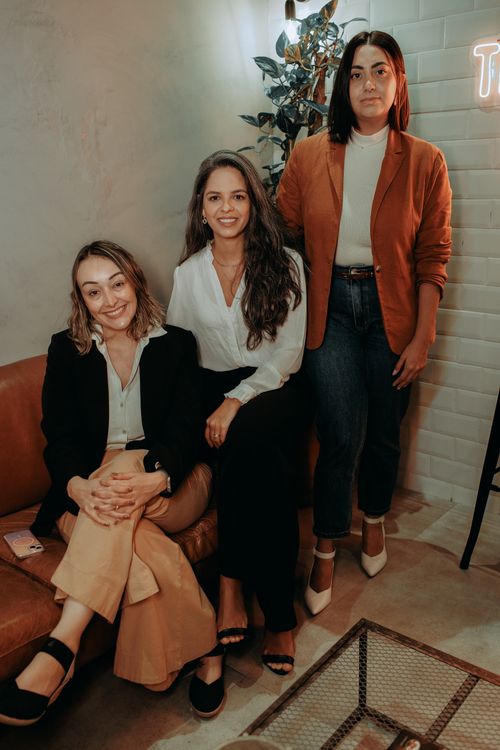 Free Three stylish women sitting and standing in a chic indoor setting, exuding confidence and elegance. Stock Photo