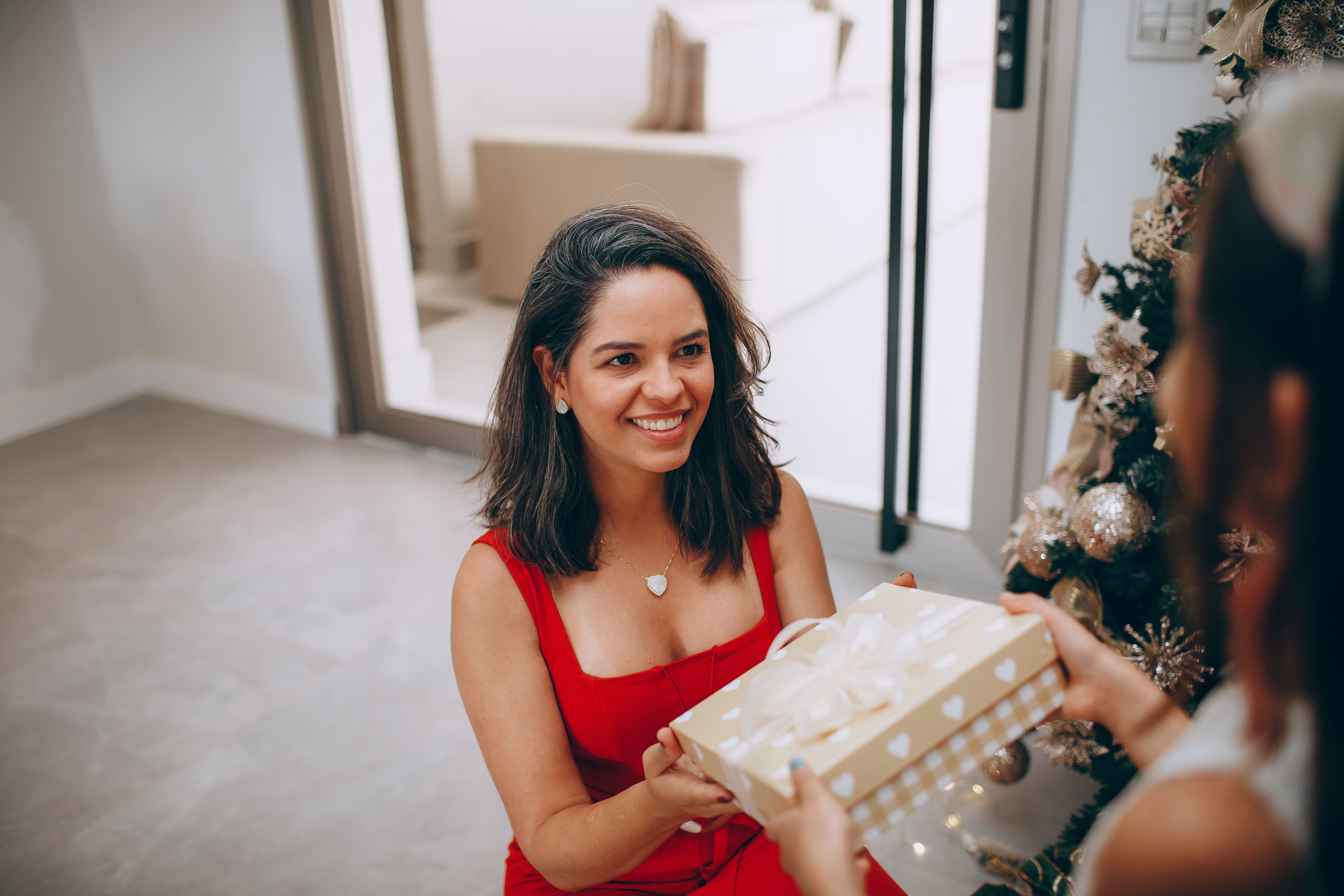 A woman in a red dress receives a Christmas gift, smiling by the festive tree indoors.