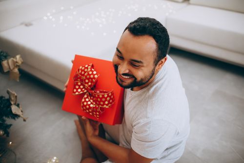 Free Joyful man with beard holding a red gift with festive bow indoors, celebrating Christmas. Stock Photo