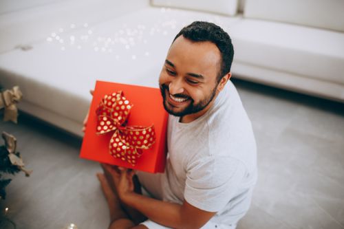 Free Smiling man holding a red Christmas gift with polka dot ribbon in a cozy living room setting. Stock Photo