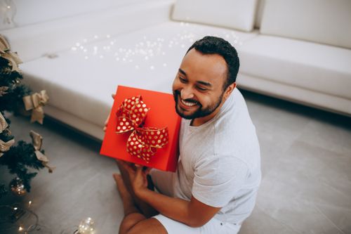 Free Smiling man with a beard holding a red Christmas present with a bow indoors. Stock Photo