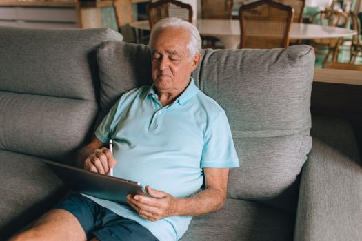 Elderly man using a tablet on a sofa, enjoying leisure time at home.