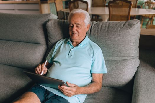 Senior man using a tablet with a stylus while relaxing on a sofa indoors.