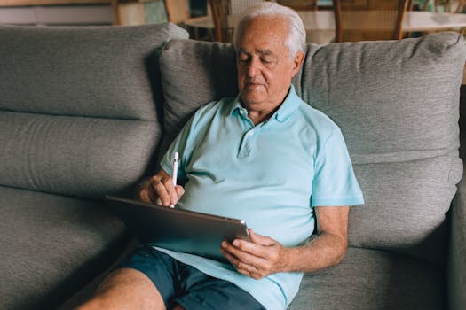 Elderly man in blue polo shirt using a tablet while lounging on a comfortable sofa.