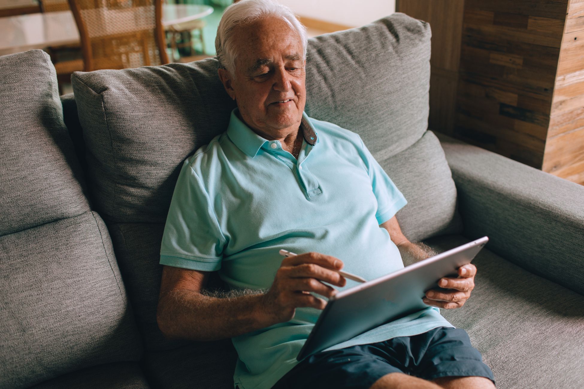 Elderly man in blue polo sitting on a couch using a tablet with stylus indoors.