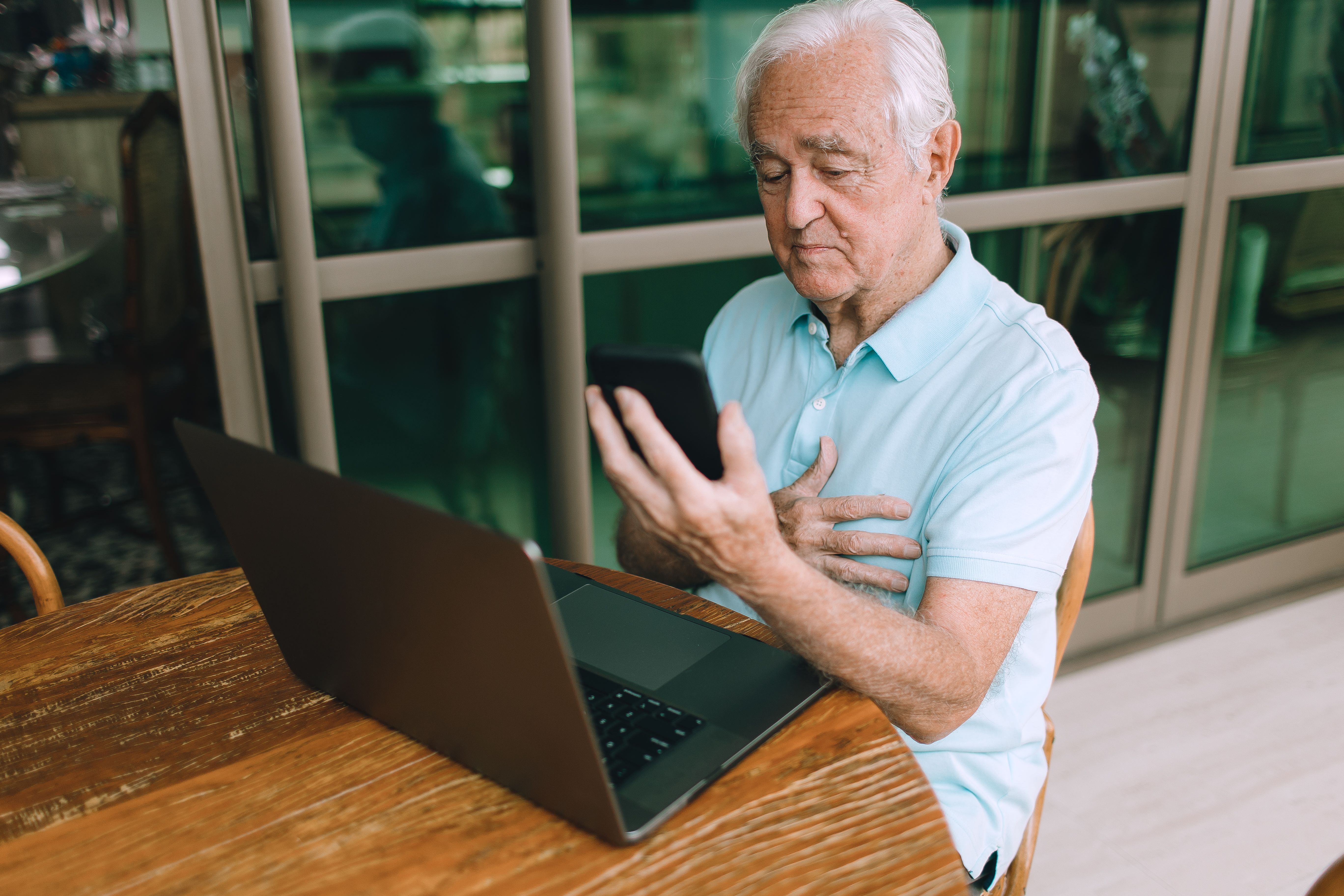 Idoso usando smartphone e laptop em uma mesa de madeira dentro de casa.