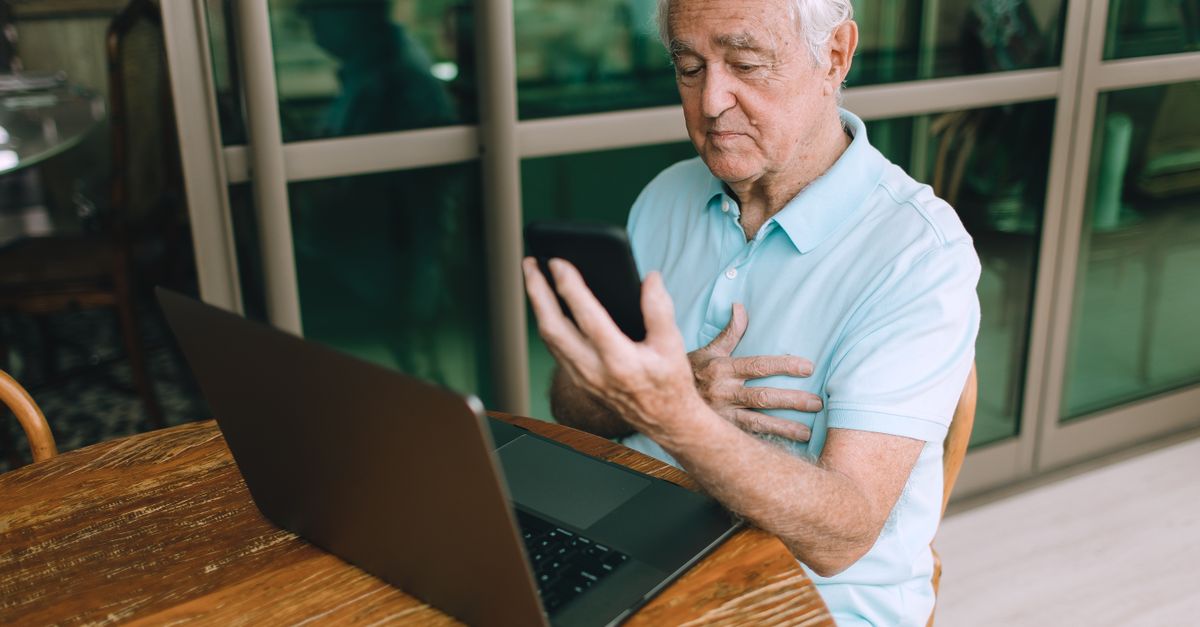 Idoso usando smartphone e laptop em uma mesa de madeira dentro de casa.