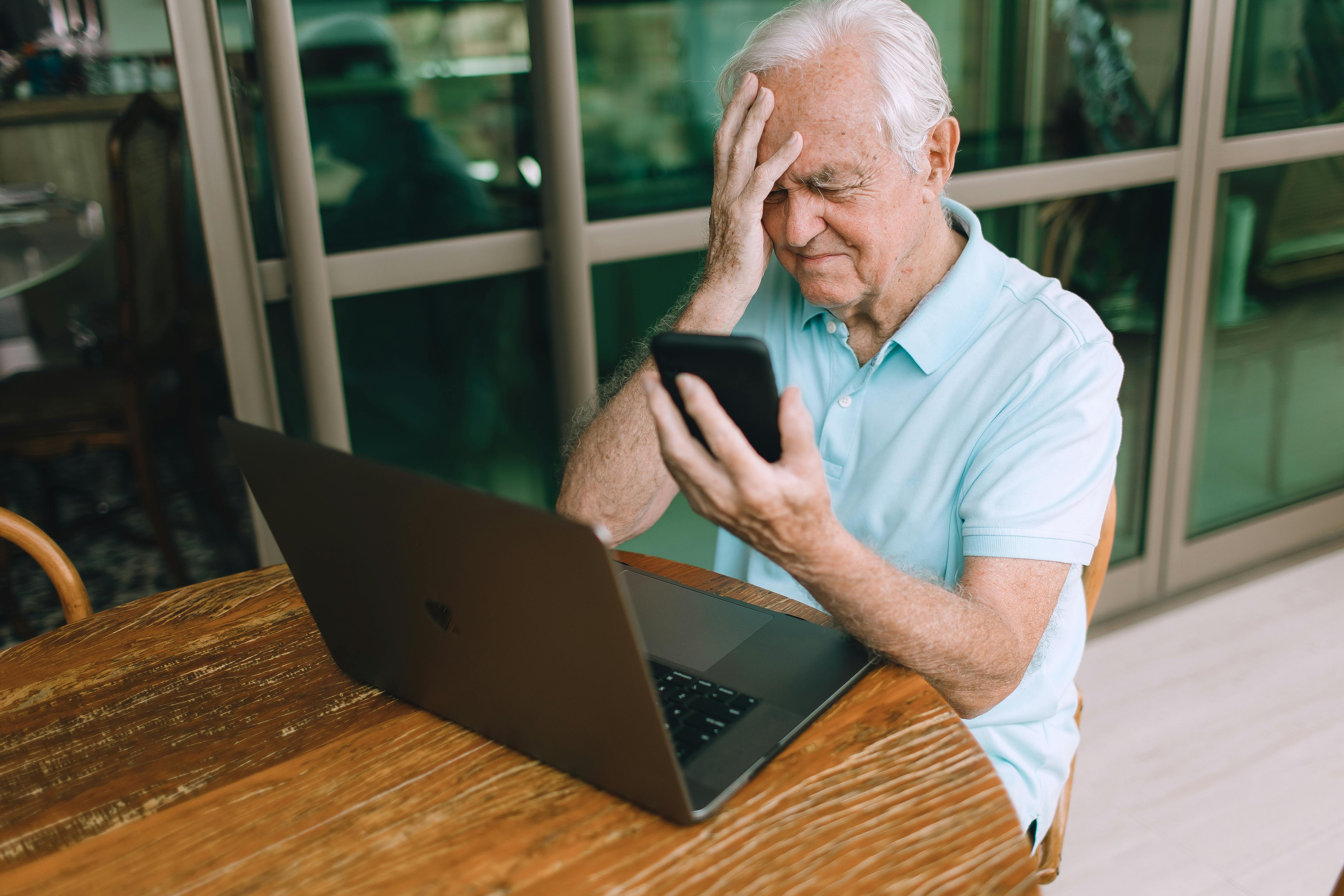 Elderly man frustrated by smartphone and laptop technology indoors at home
