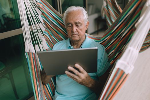 Elderly man comfortably using a tablet in a colorful hammock indoors.
