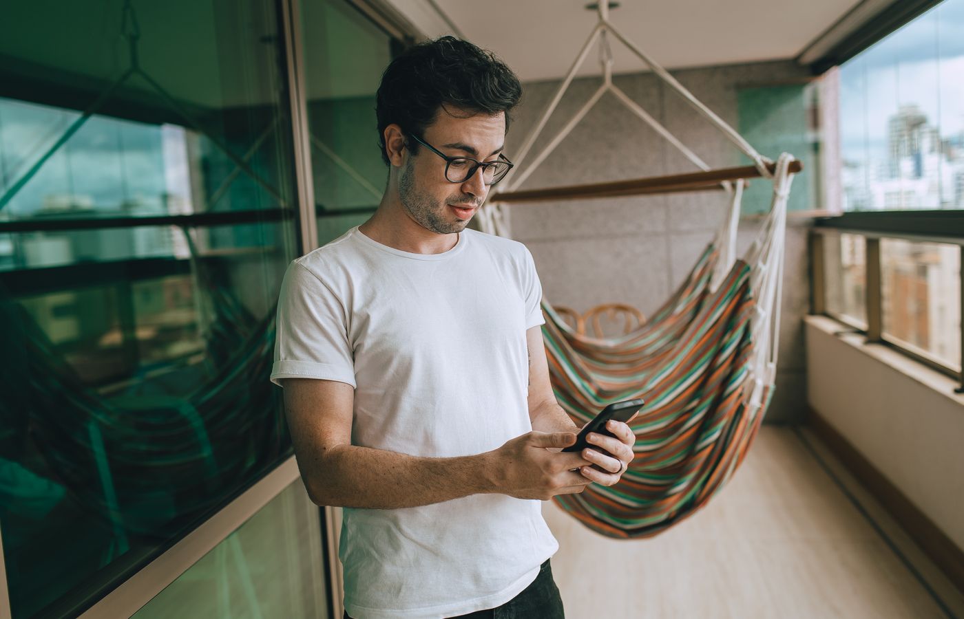 Young man in glasses using smartphone on his balcony with a hammock in the background.