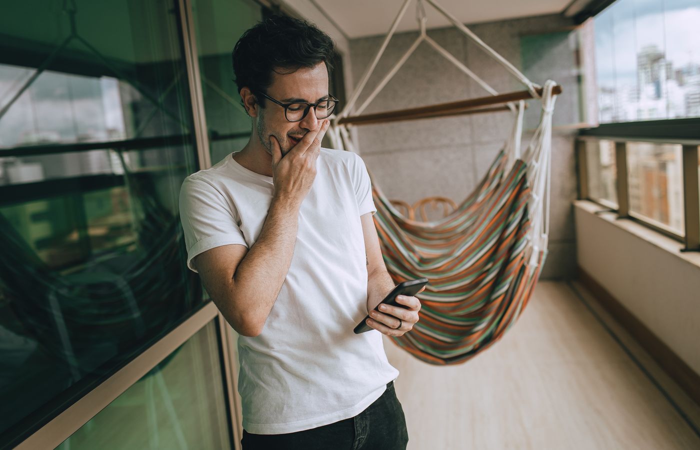 Casual portrait of a man on a balcony, using a smartphone, wearing glasses and a white t-shirt.