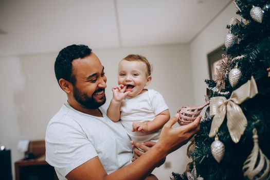 A joyful father and baby decorate a festive Christmas tree indoors, sharing a precious moment.