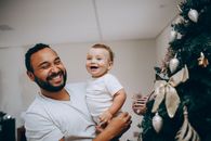 A father and his baby son smiling happily by a festive Christmas tree indoors.