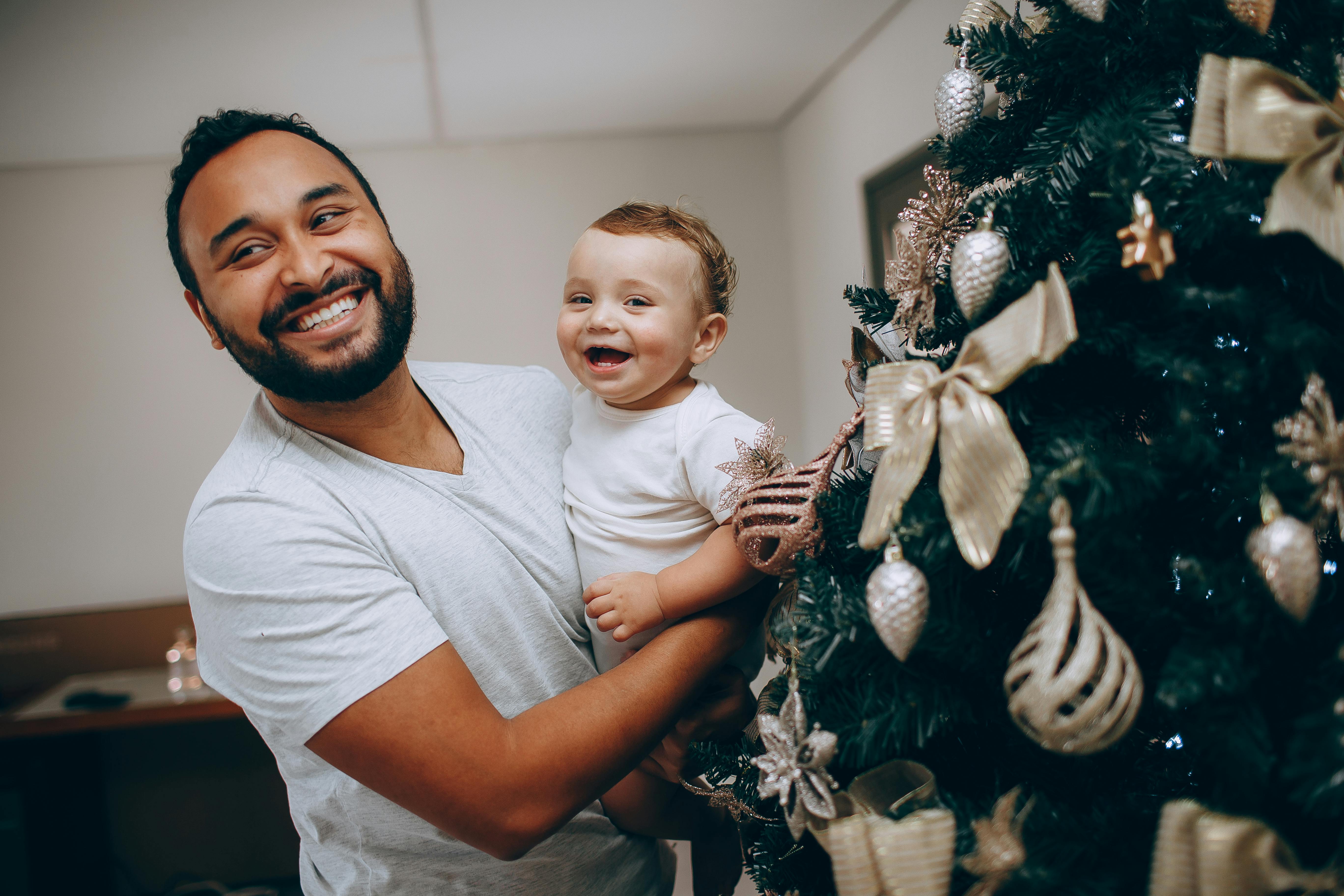 A smiling father holds his cheerful son near a beautifully decorated Christmas tree indoors.