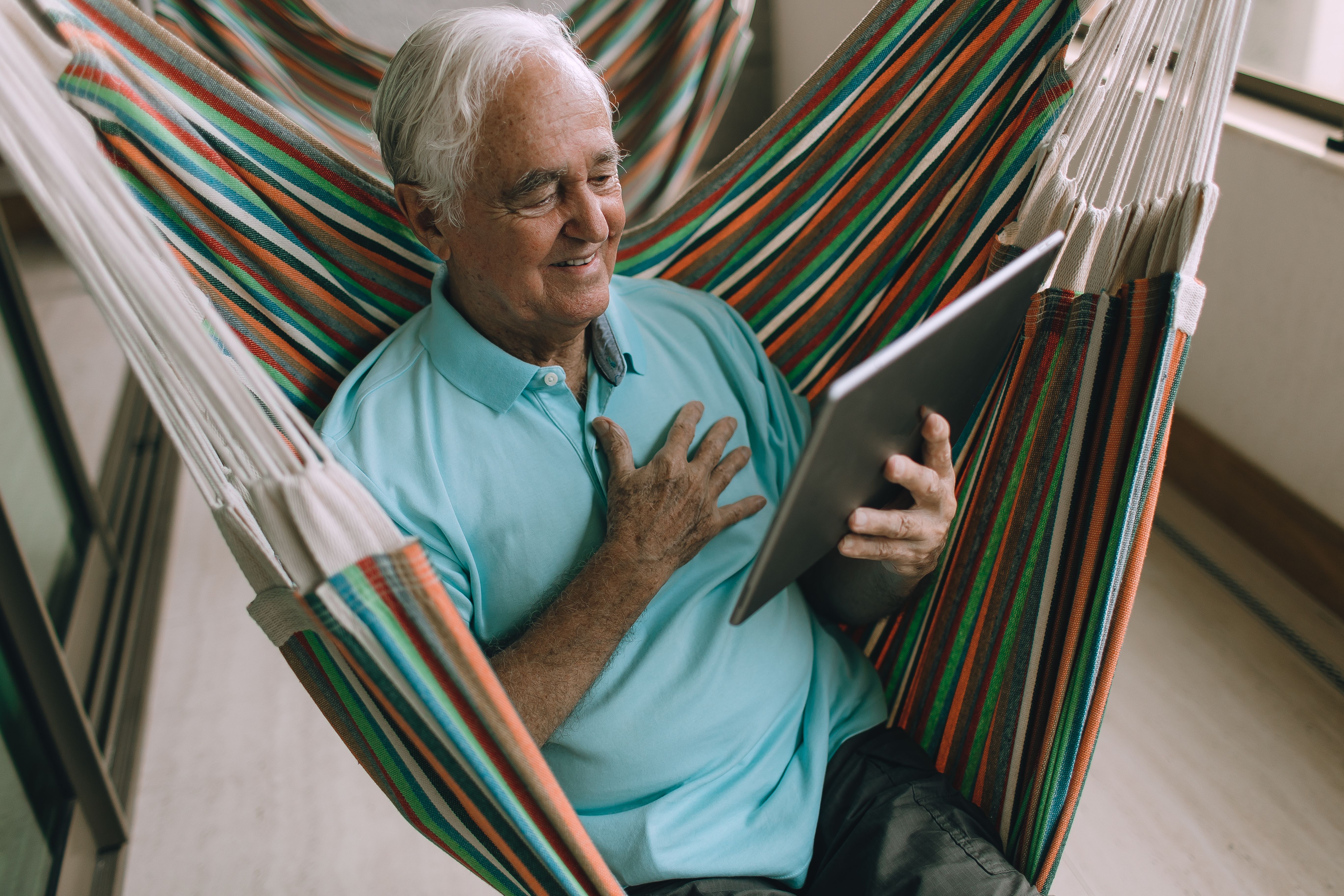 Elderly man enjoying leisure time on a hammock while using a tablet indoors.