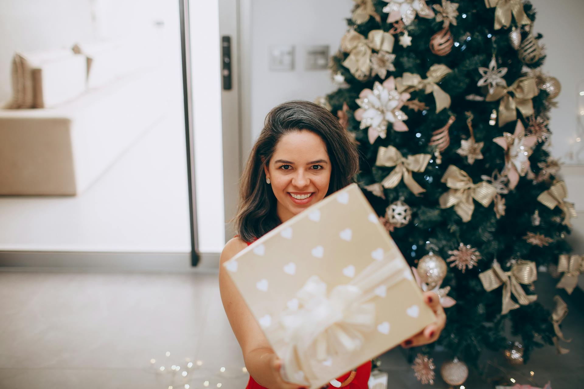 Smiling woman offering a beautifully wrapped present in front of a decorated Christmas tree.