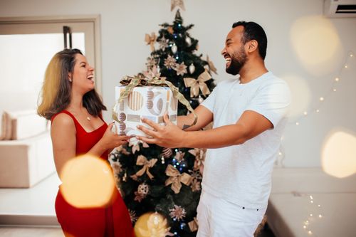 Free A couple joyfully exchanges gifts by a decorated Christmas tree indoors. Stock Photo