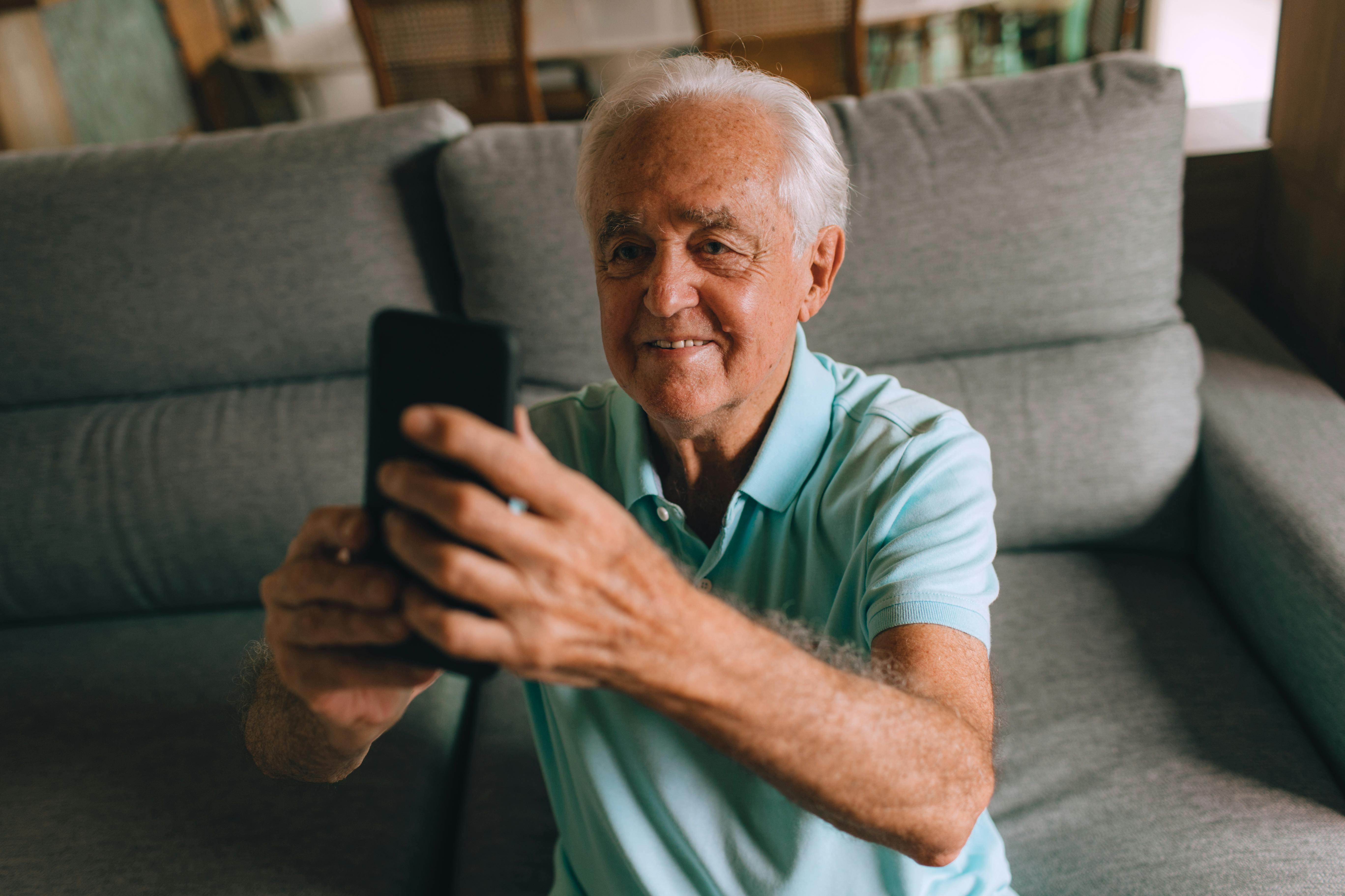 Elderly man in a living room using a smartphone, wearing a blue polo shirt.