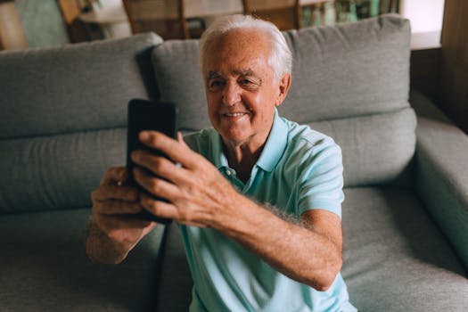 Senior man smiling while using a smartphone on a sofa indoors, expressing joy and engagement.