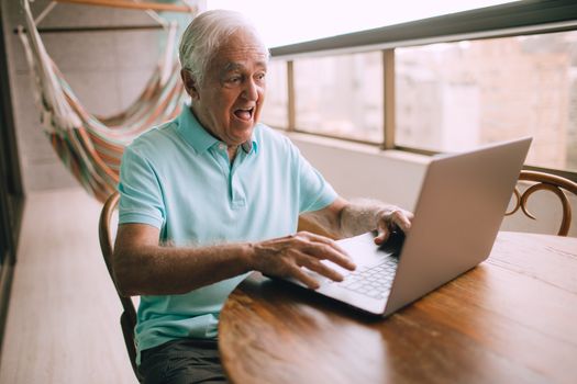 Senior adult sitting indoors, surprised, using a laptop at a wooden table.