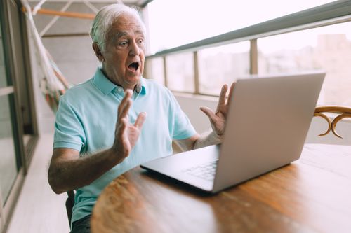 Free Senior man in blue polo shirt surprised by content on laptop while sitting on a balcony. Stock Photo