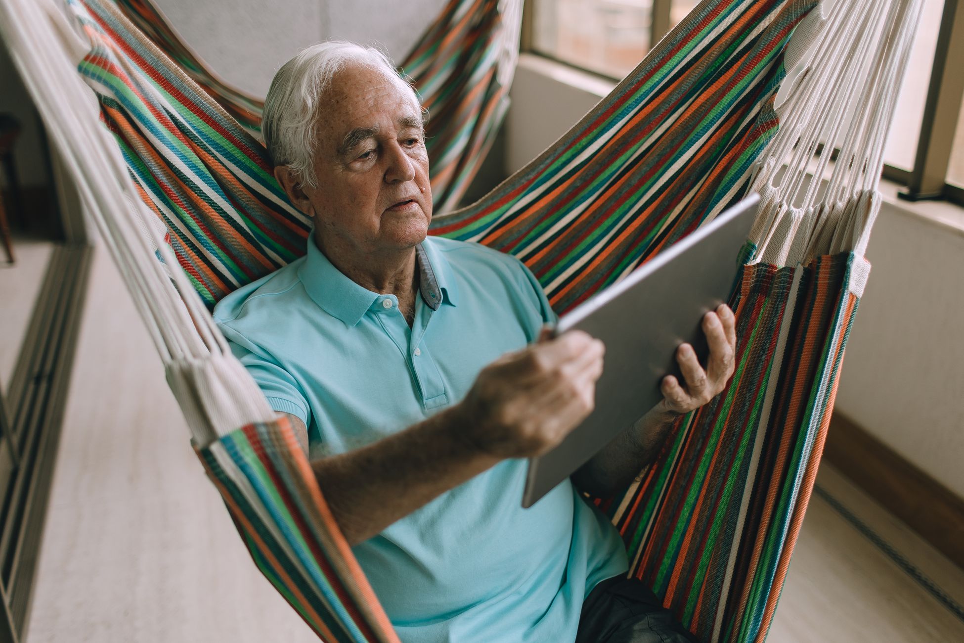 Elderly man using a tablet while relaxing on a hammock on a balcony.