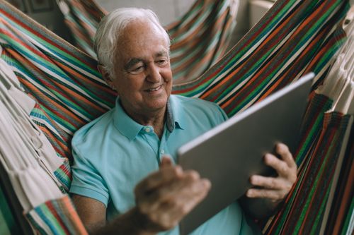 Free Senior man enjoys leisure time using a tablet in a colorful hammock. Stock Photo