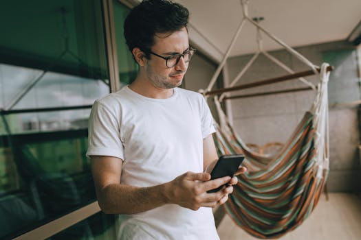 Portrait of a young man with glasses using smartphone near a colorful hammock on a balcony.
