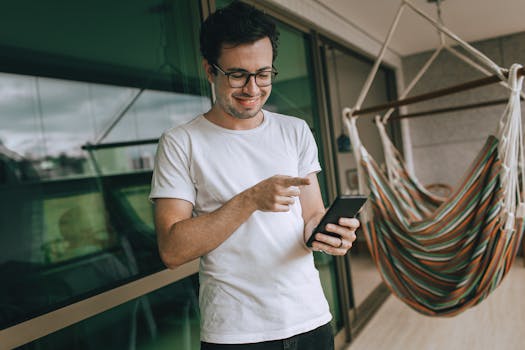 A smiling young man using a smartphone on a balcony next to a colorful hammock.