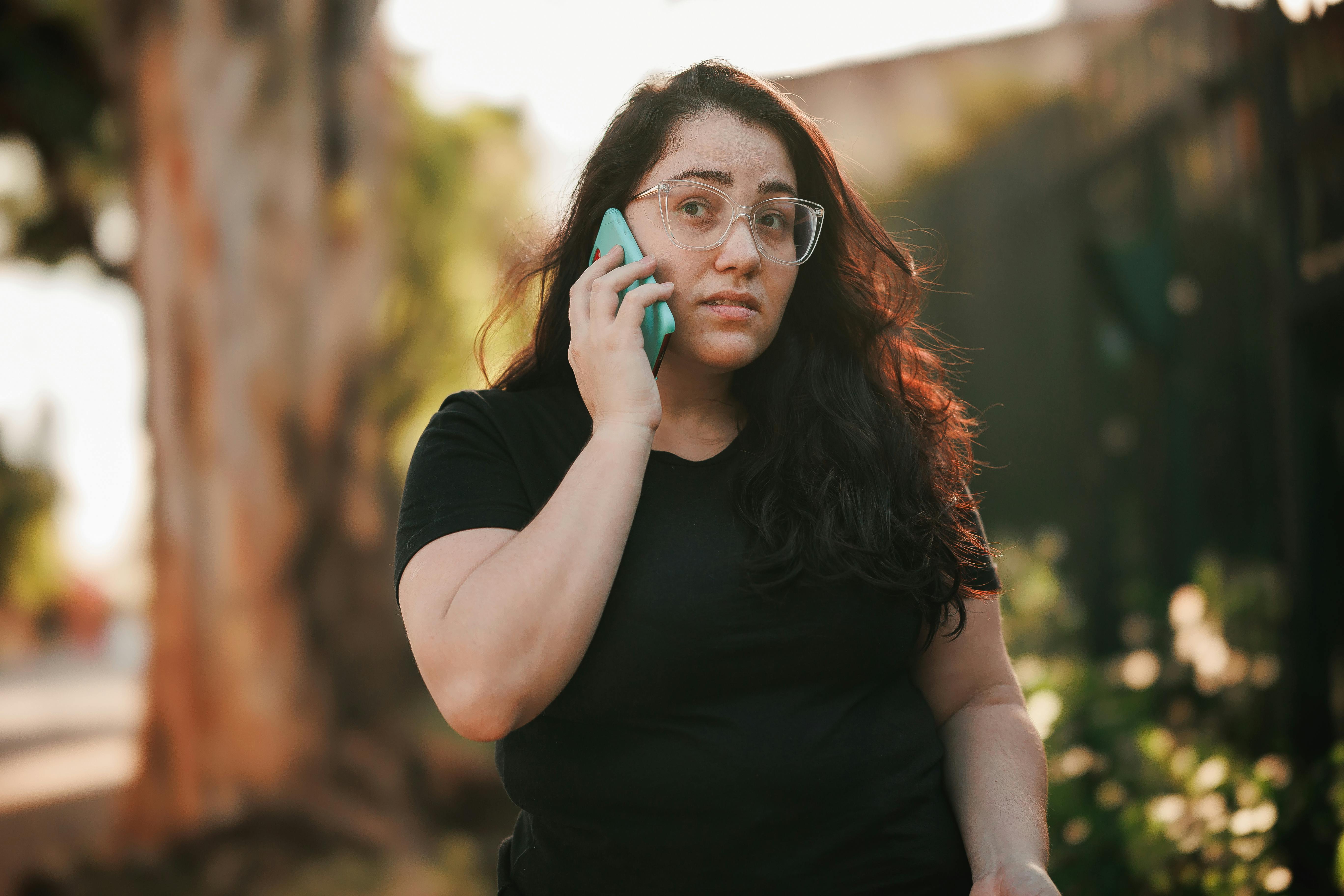 Woman with eyeglasses talks on smartphone in sunny park setting.