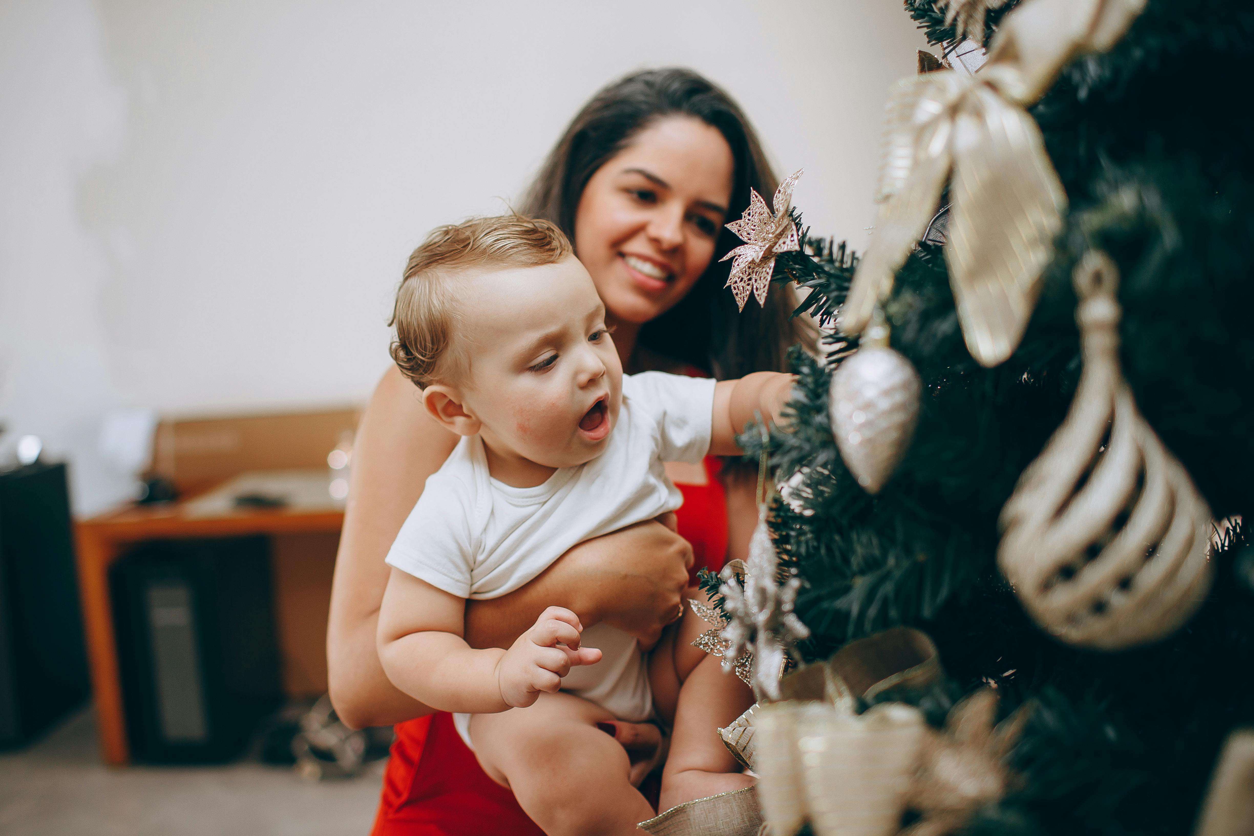 Smiling mother helps baby decorate a Christmas tree inside their home, capturing holiday joy.
