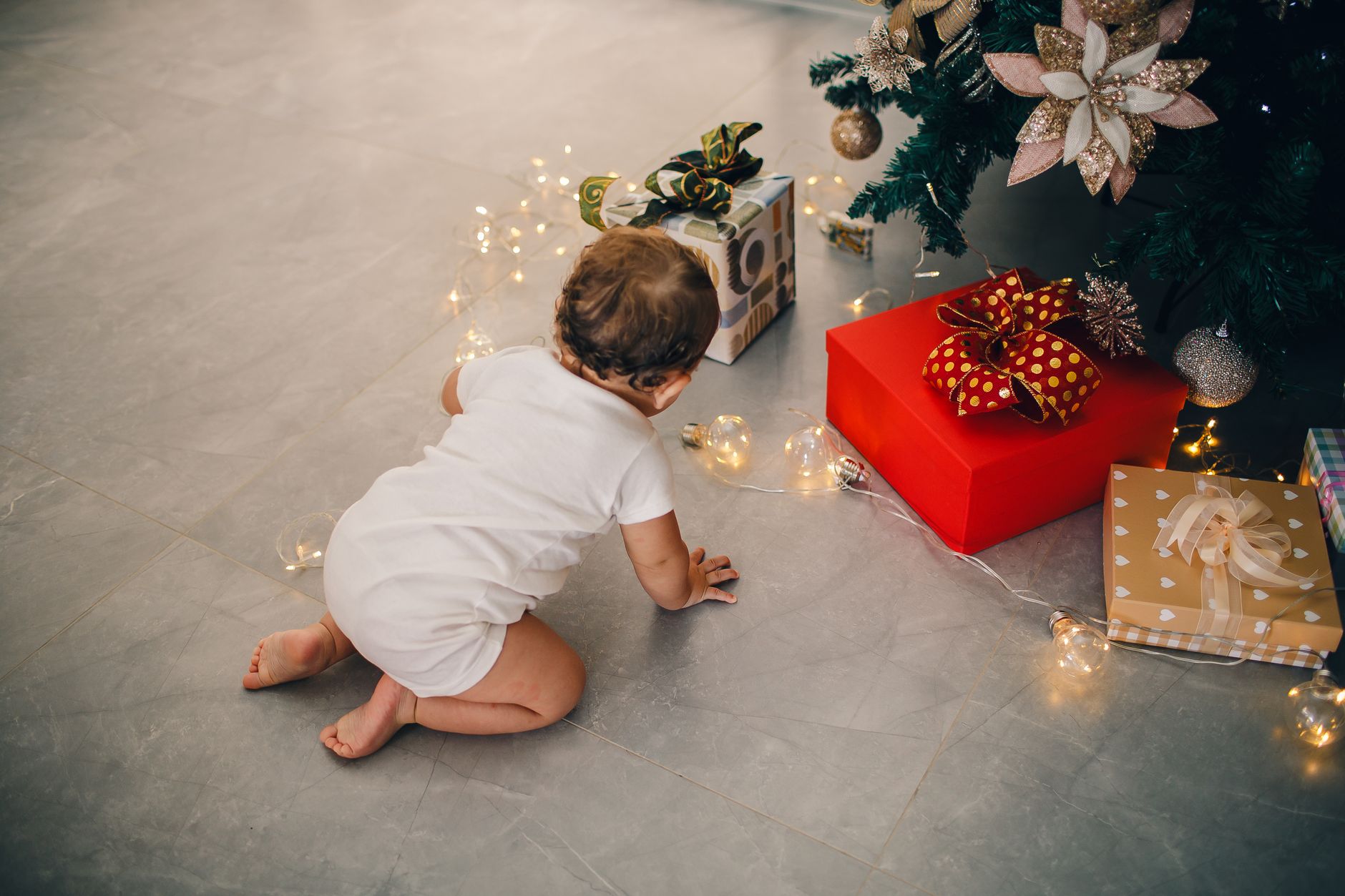 Cute baby in white onesie crawls near a decorated Christmas tree with presents and lights.