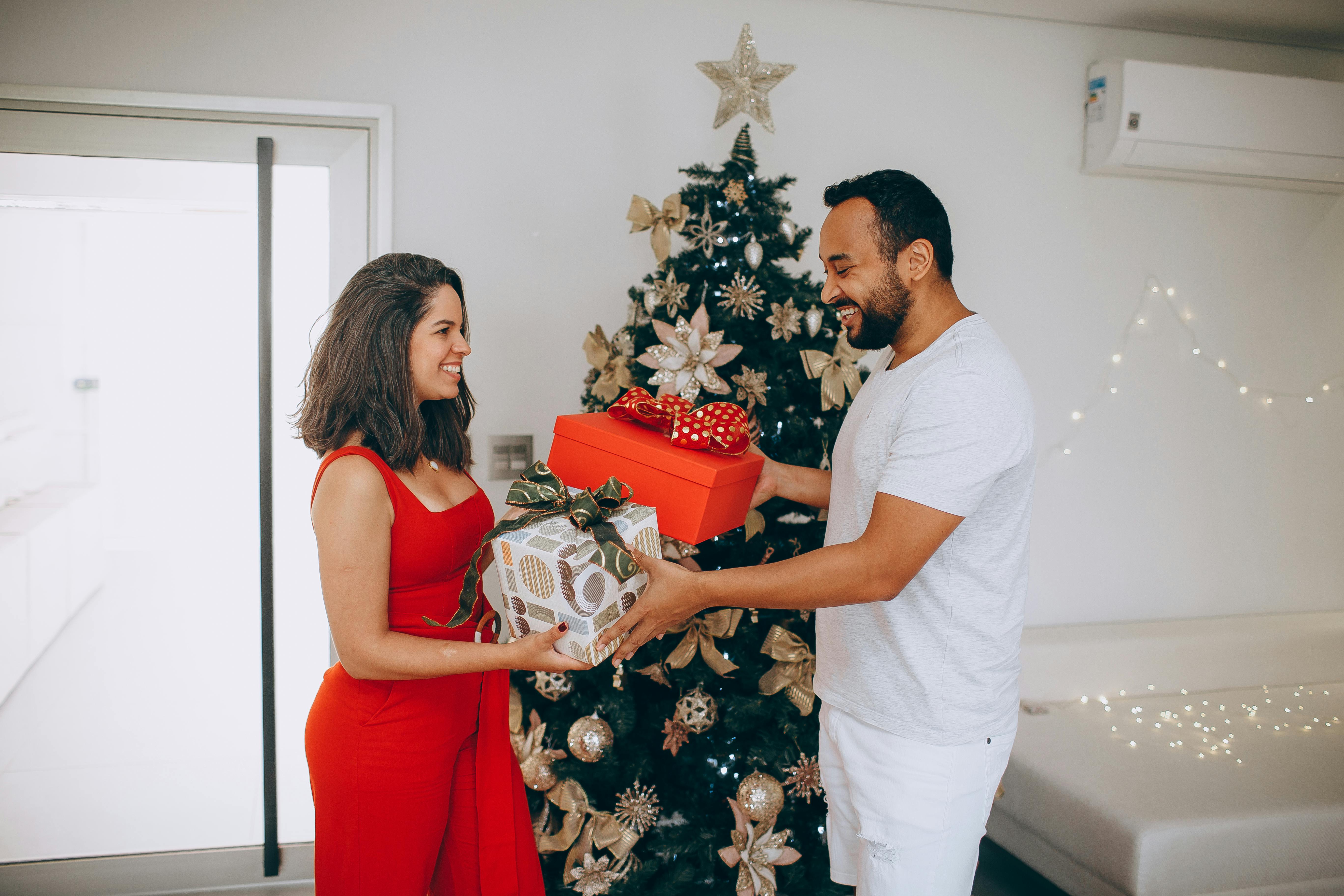 A joyful couple exchanging gifts beside a decorated Christmas tree inside a cozy room.