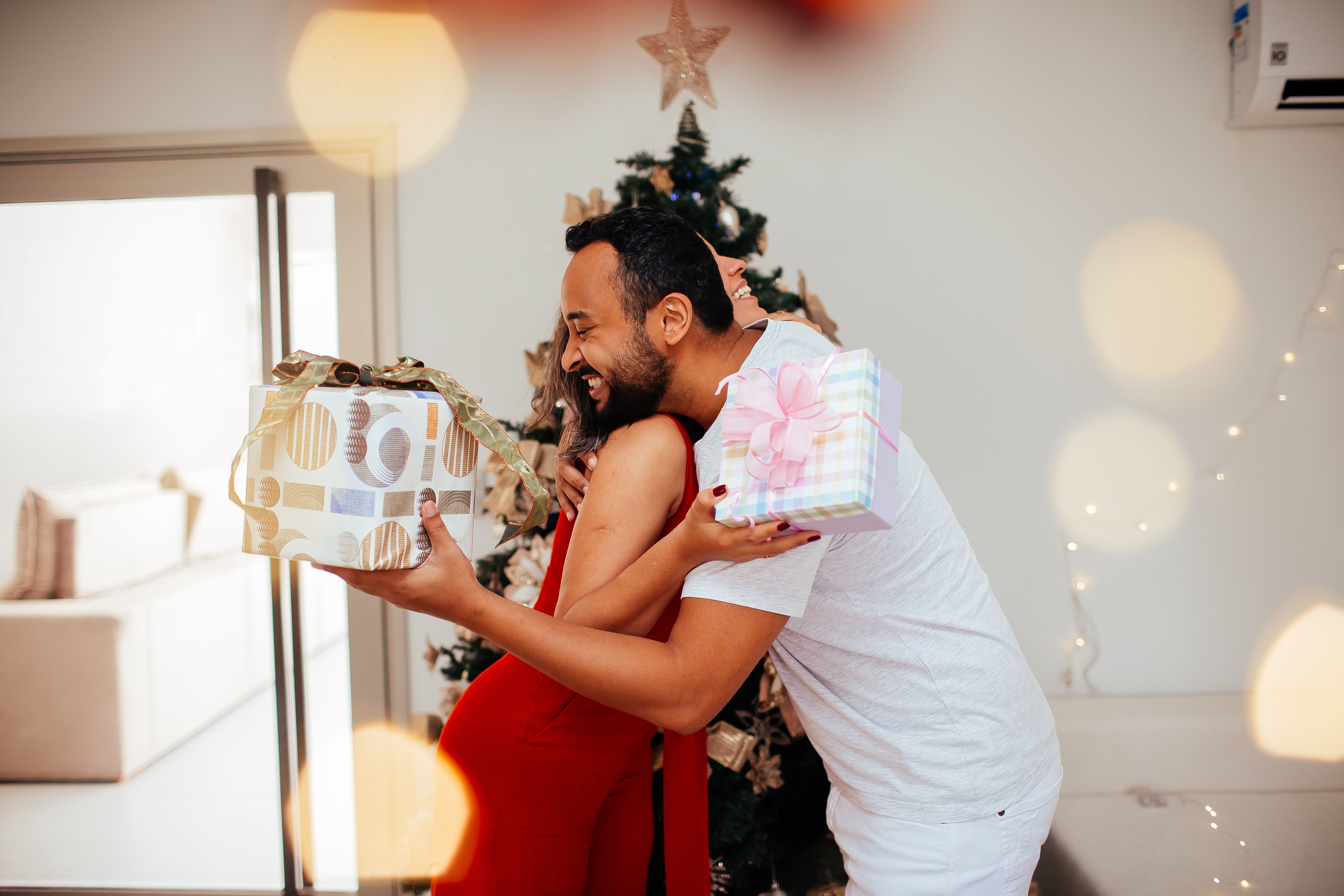A couple joyfully exchanges gifts near a decorated Christmas tree, creating a warm holiday atmosphere.