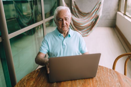 Elderly man sitting at a wooden table, using a laptop in a bright indoor setting, near a balcony.