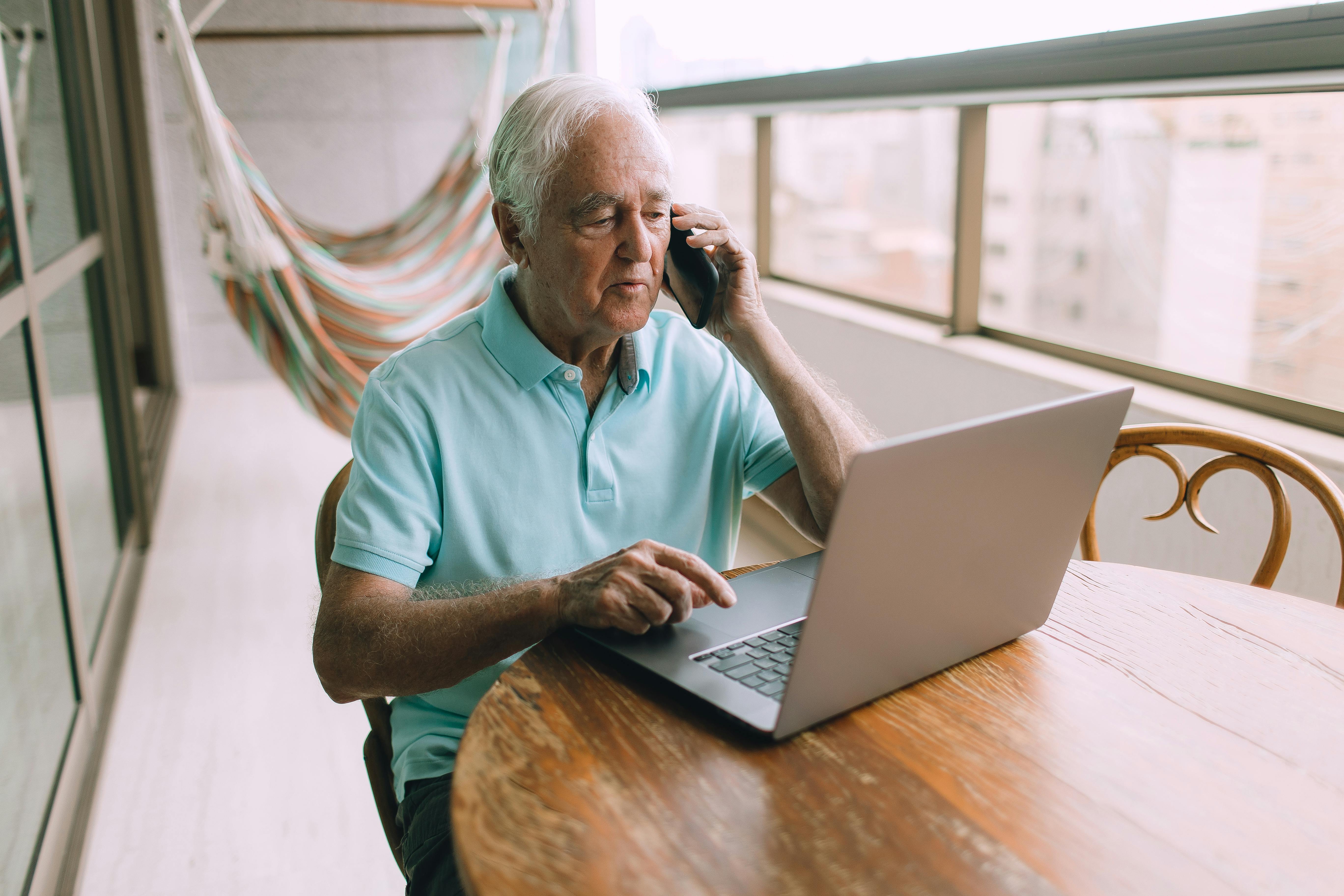 Elderly man multitasking with a phone call and laptop on a balcony.