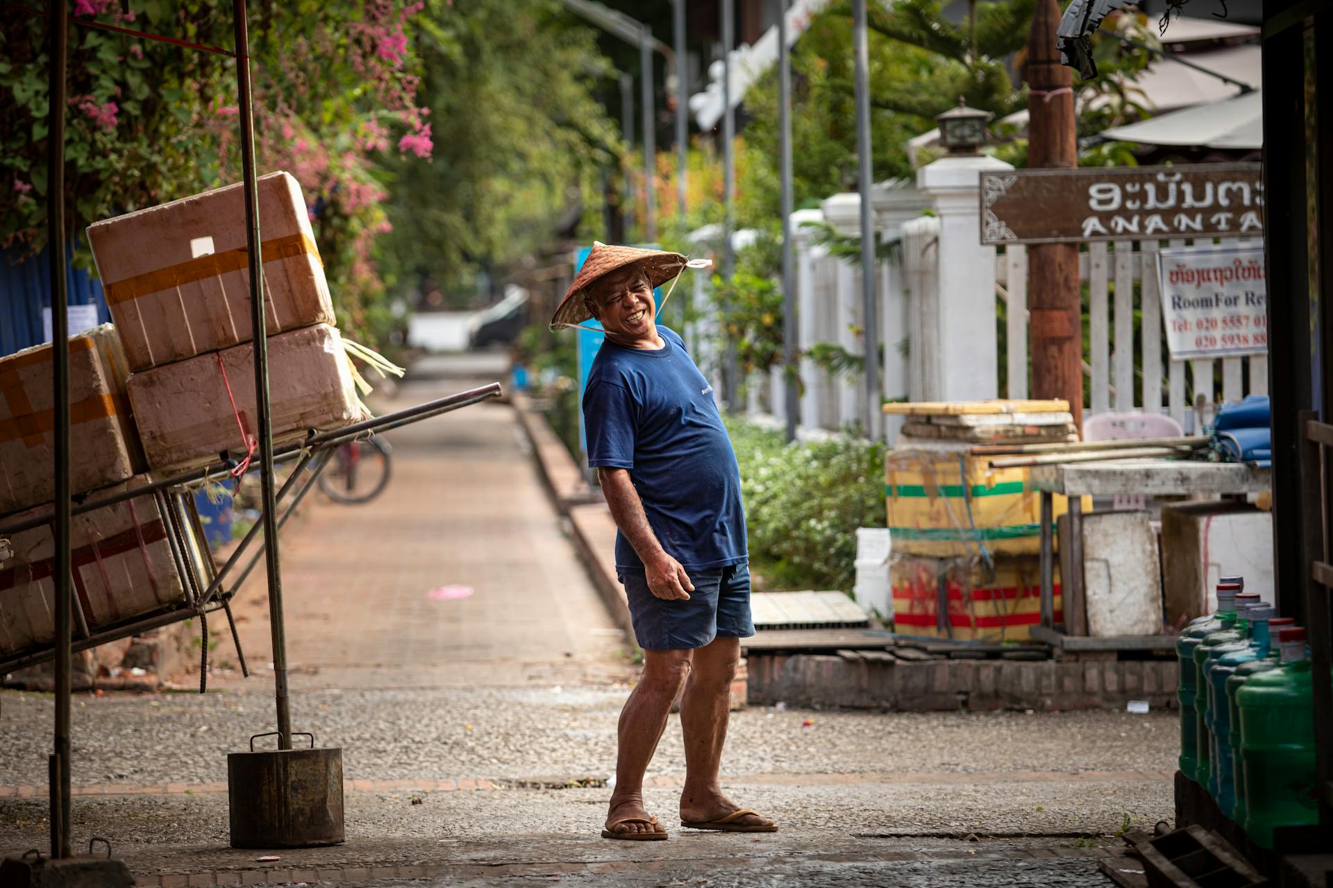 A street vendor stands in a vibrant Southeast Asian market with colorful boxes and a sunny atmosphere.