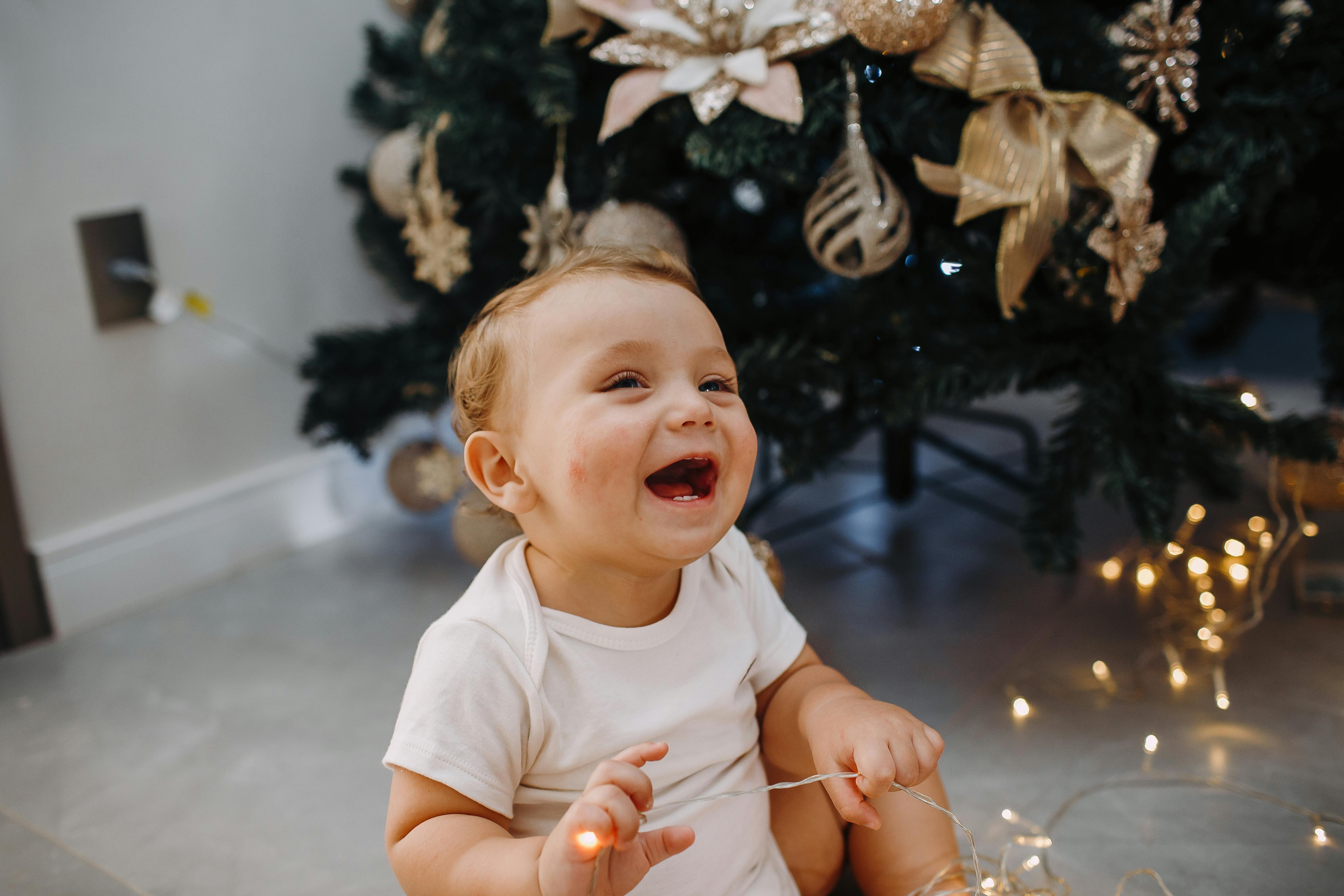A smiling baby playing with lights under a beautifully decorated Christmas tree indoors.