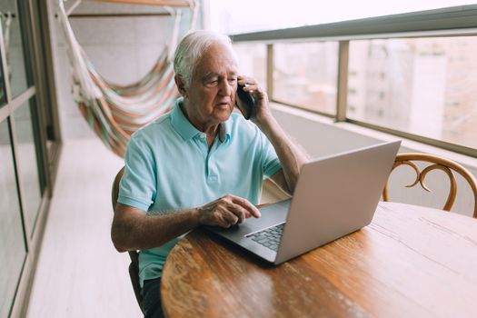 Senior man multitasking with a laptop and smartphone on a bright indoor balcony, focused on communication.