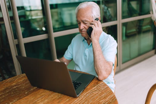 Senior man with gray hair multitasking with laptop and phone indoors.