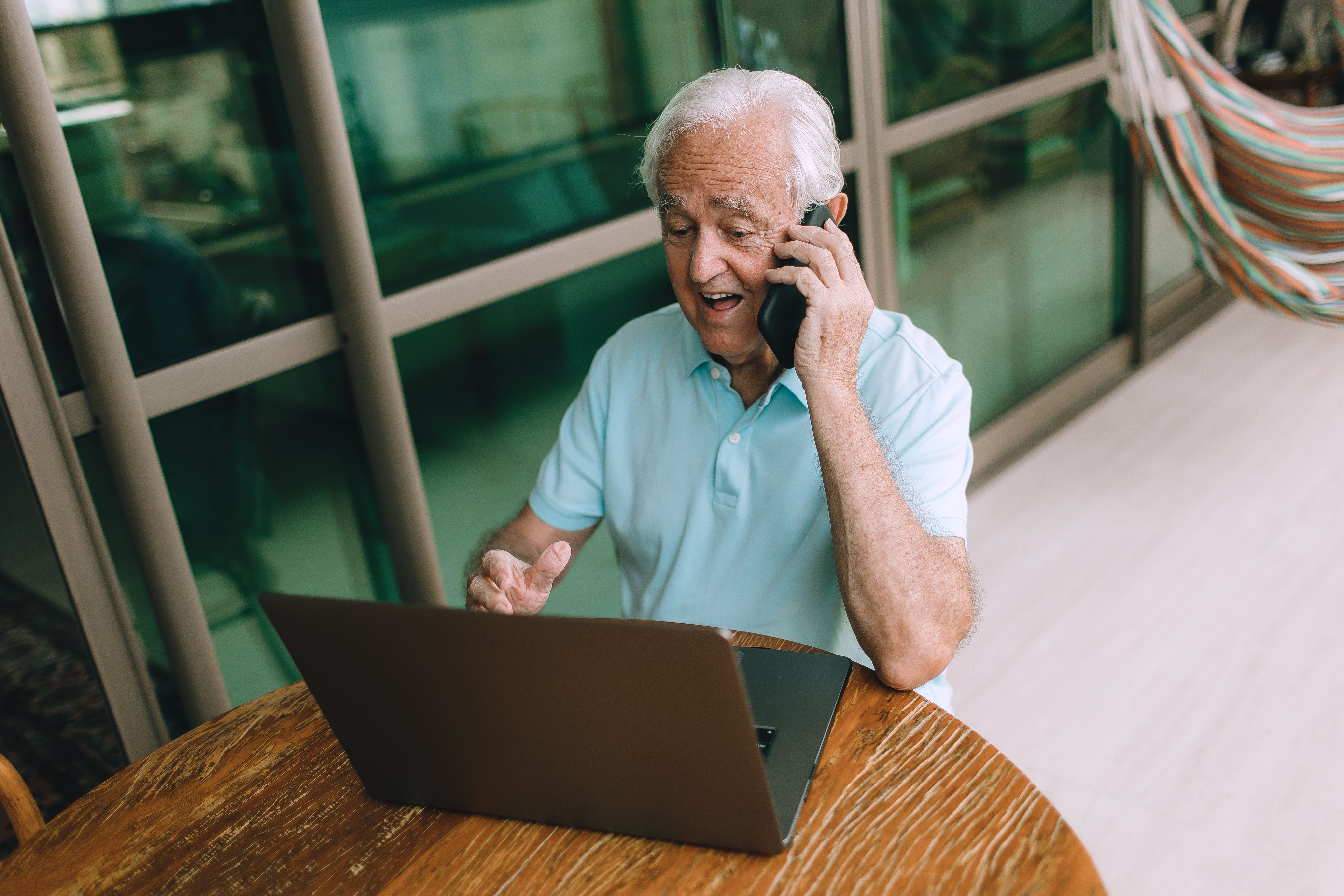 Elderly man multitasking on phone and laptop indoors with modern decor.