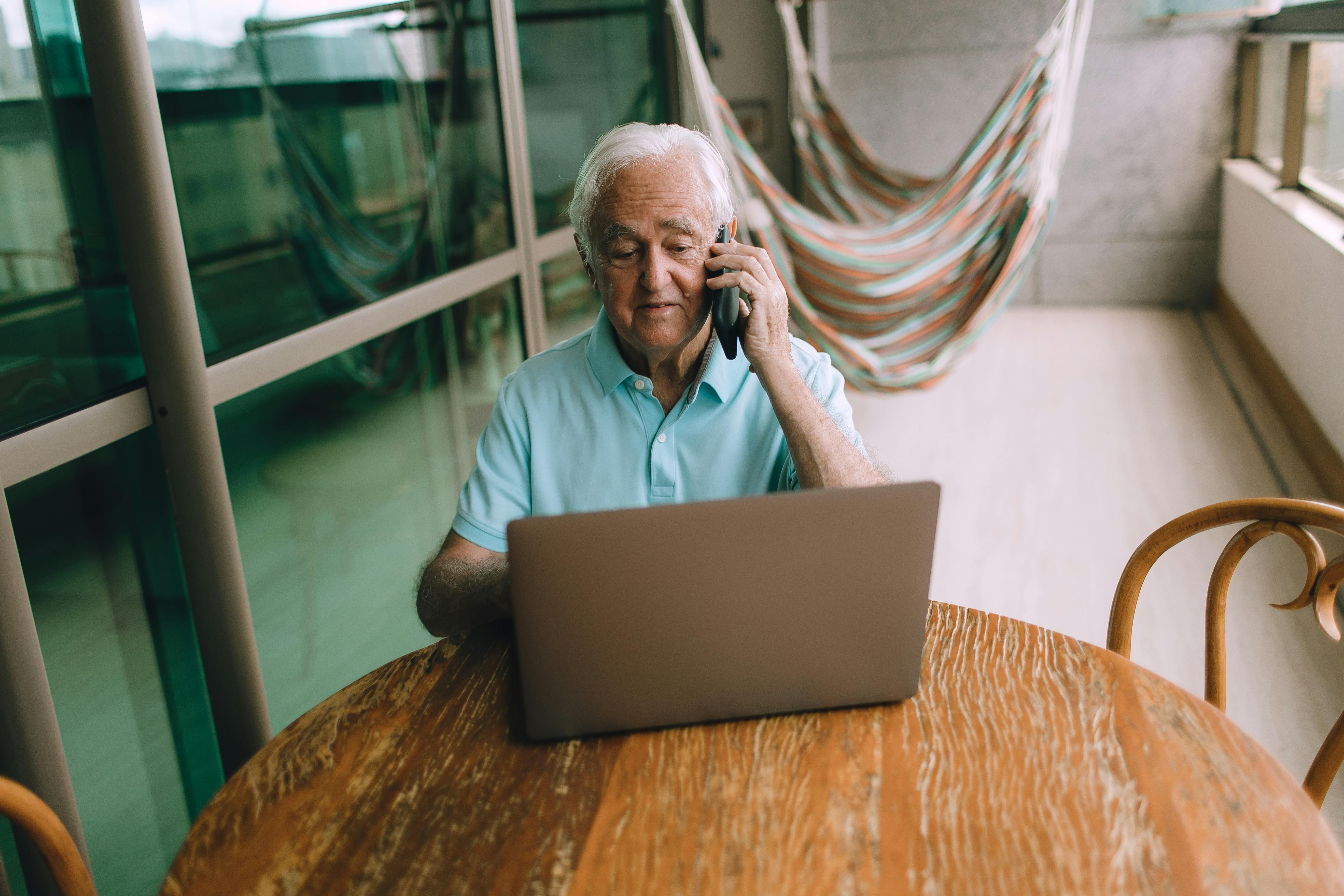 Elderly man multitasking on laptop and phone, sitting at a wooden table indoors with hammocks in the background.