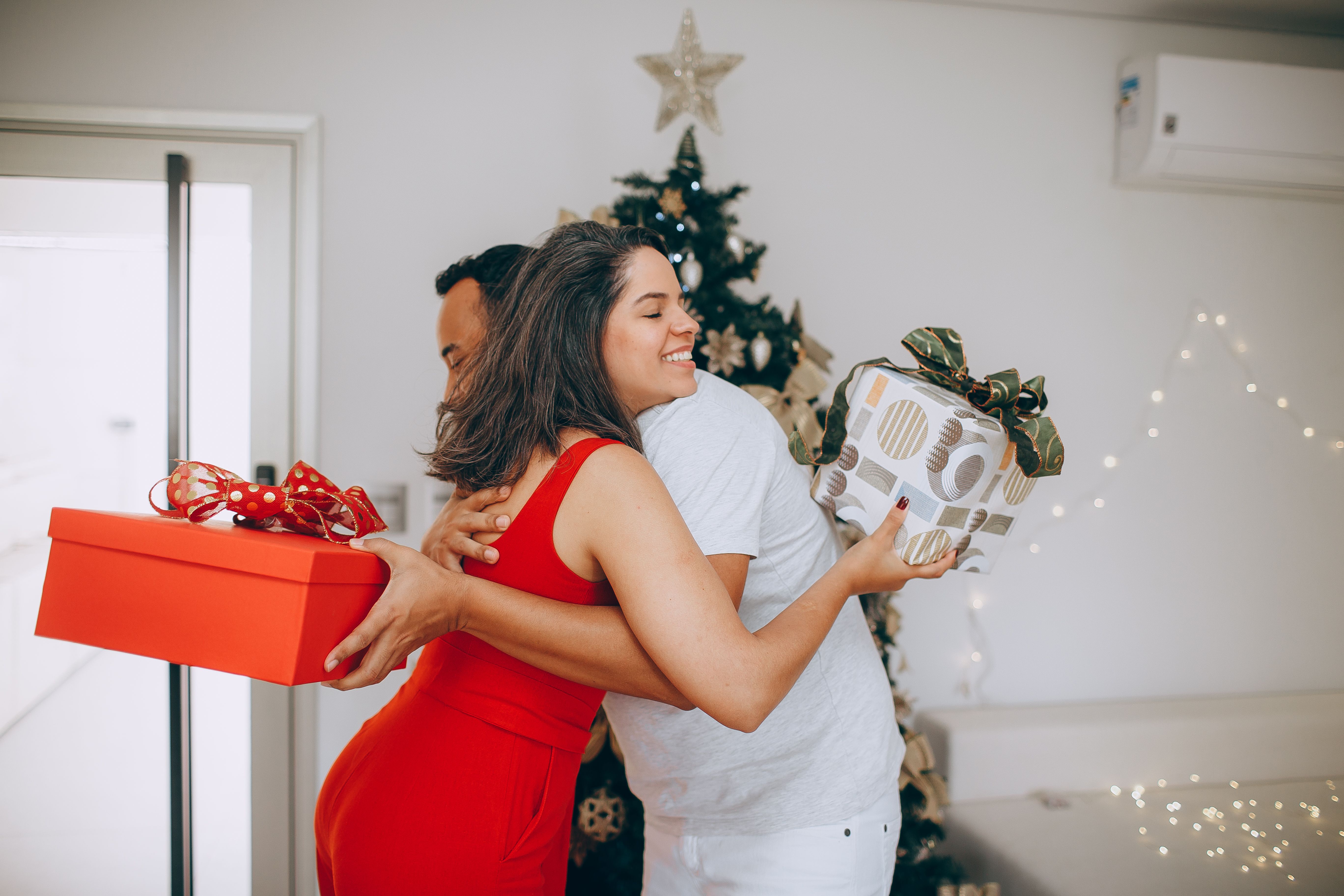 Two adults exchanging gifts in front of a decorated Christmas tree, embracing with happiness.