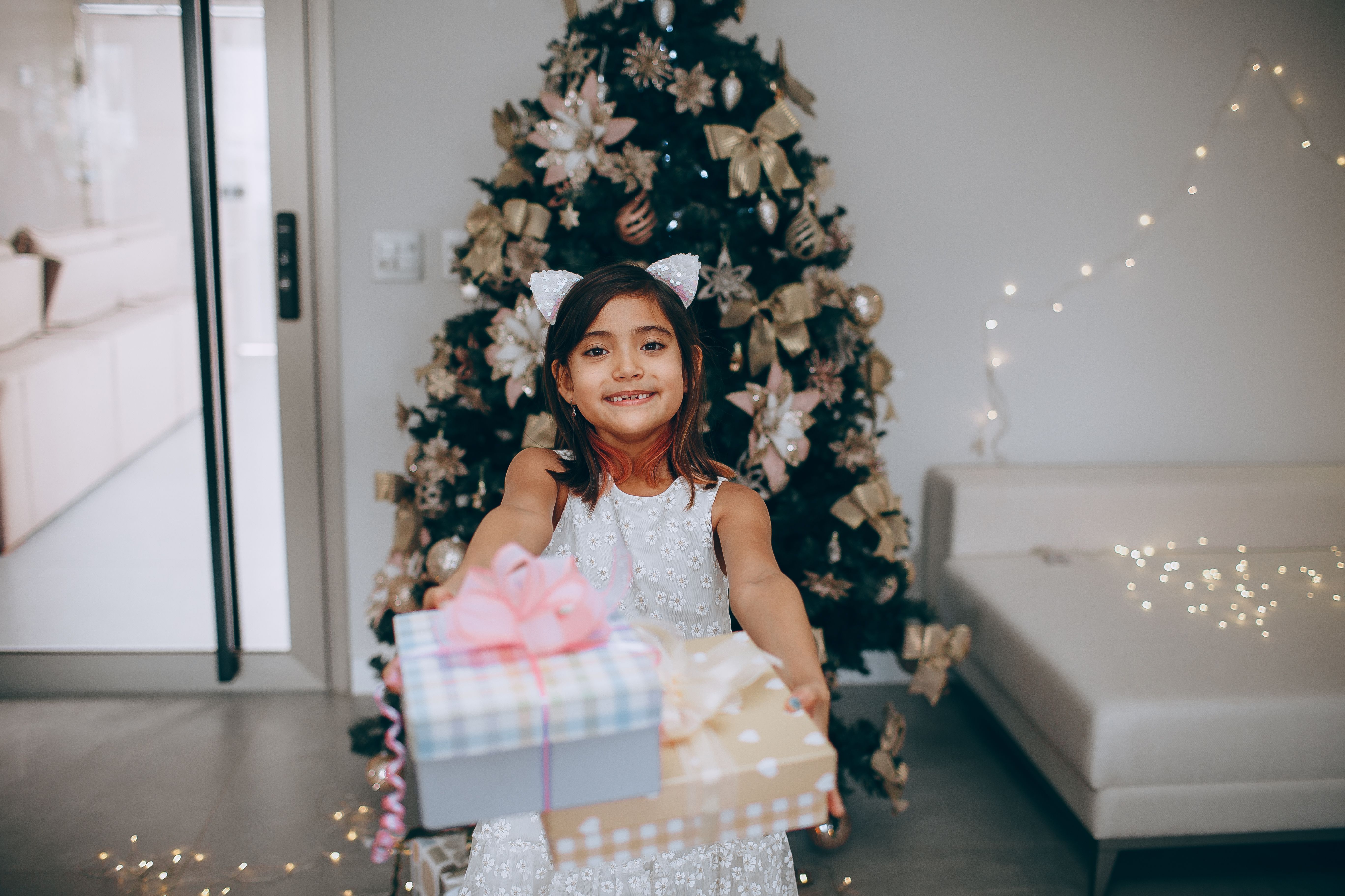 Delighted girl in festive attire offering gifts in a Christmas-decorated living room.