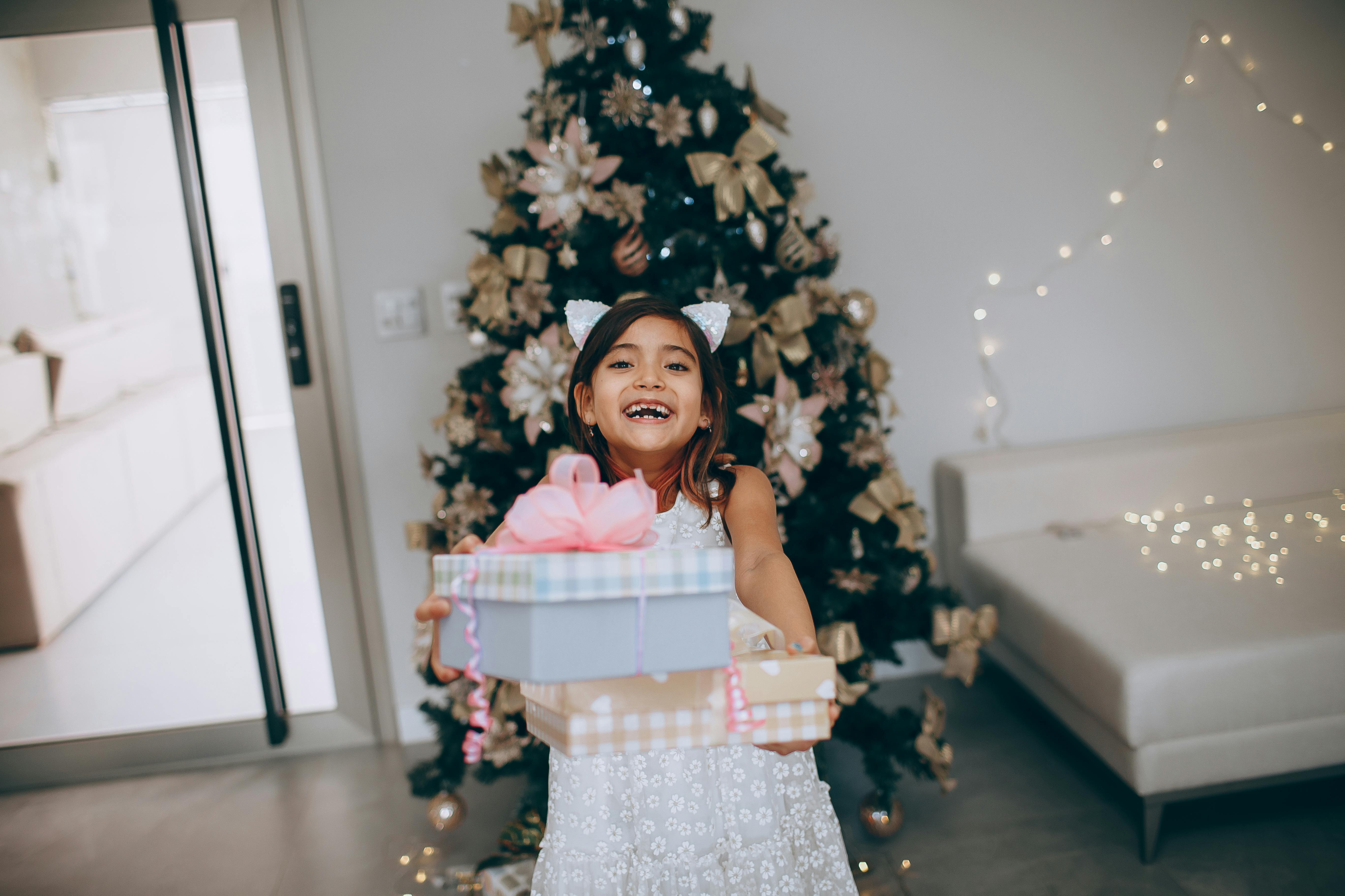 Smiling child holding presents in front of a beautifully decorated Christmas tree indoors.