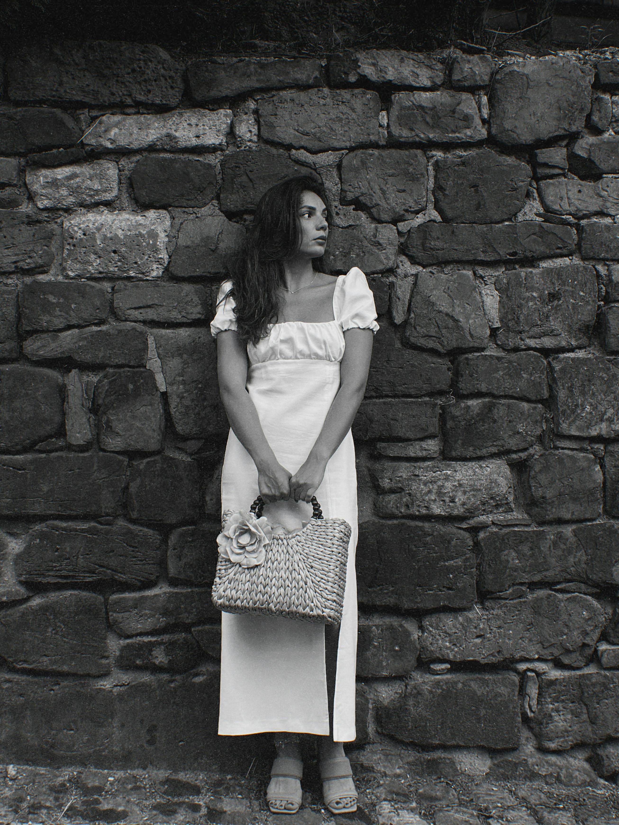 Black and white photo of a woman in a white dress standing by a stone wall.