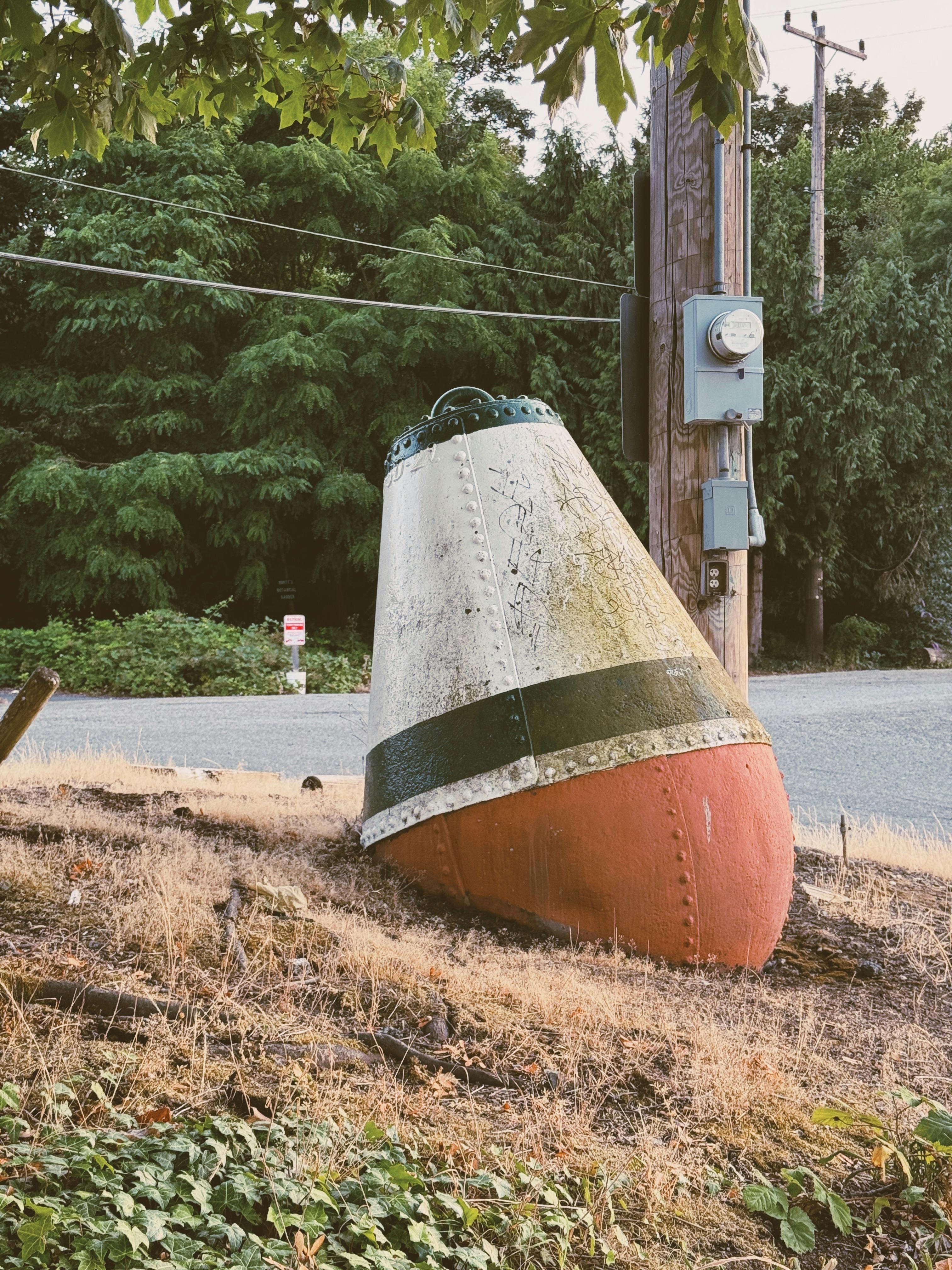 Weathered buoy resting on dry ground by a rural road surrounded by greenery.