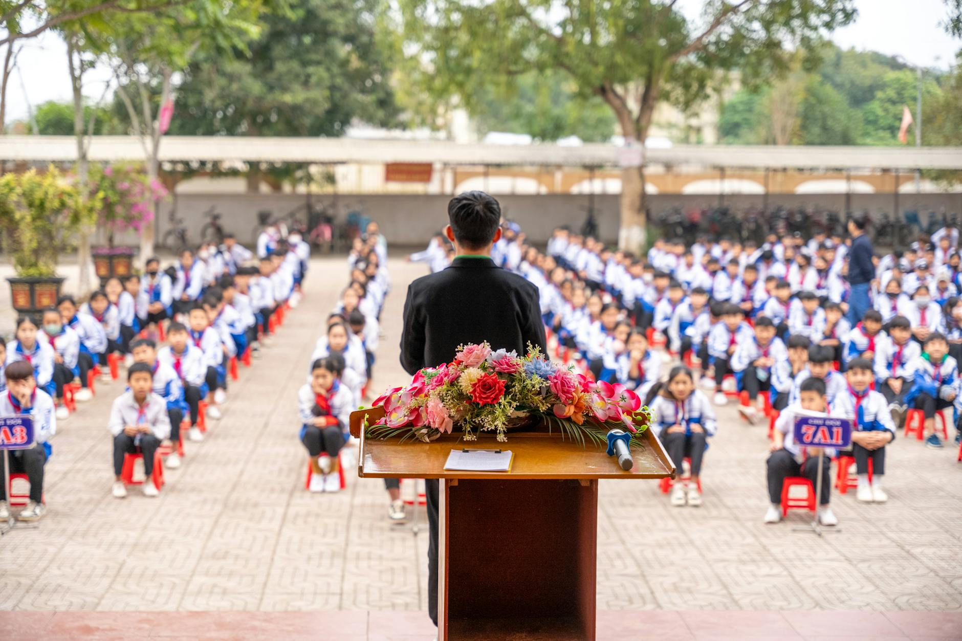 A teacher addresses students seated in an outdoor assembly at a school.